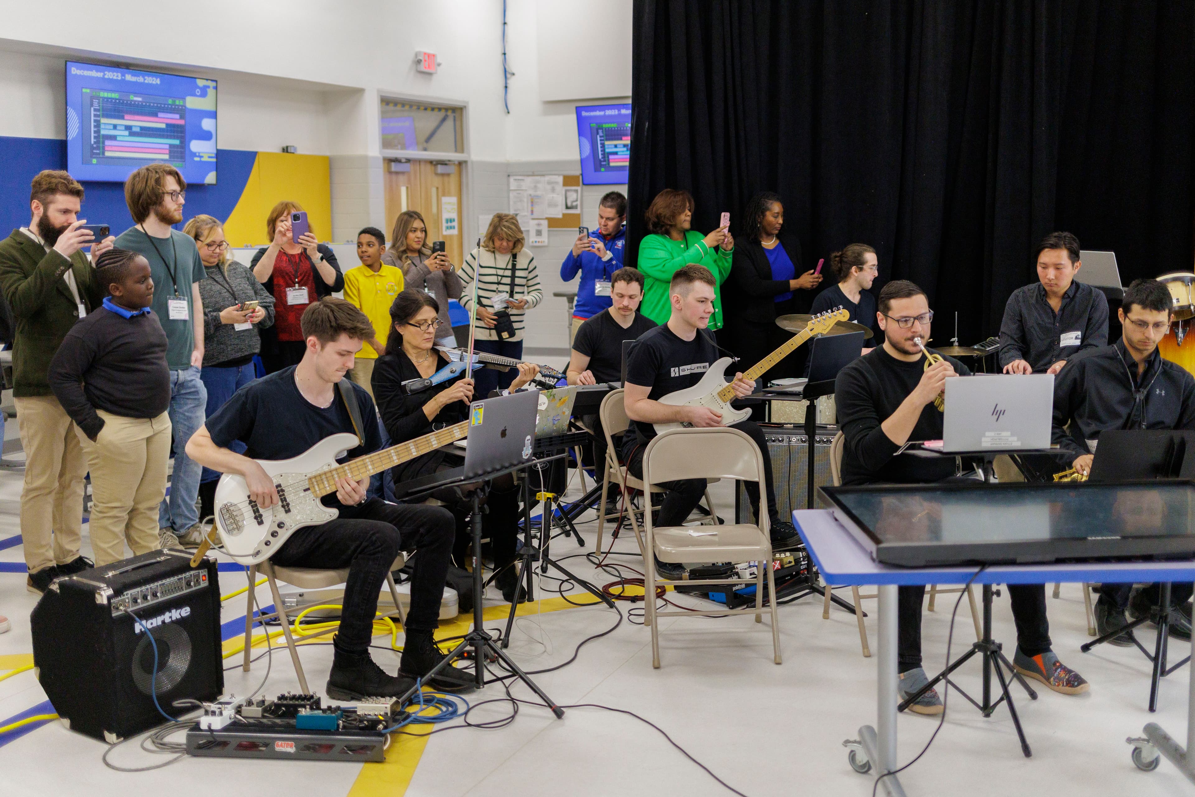 A full band performs student compositions in a school gym while families record from the back of the room