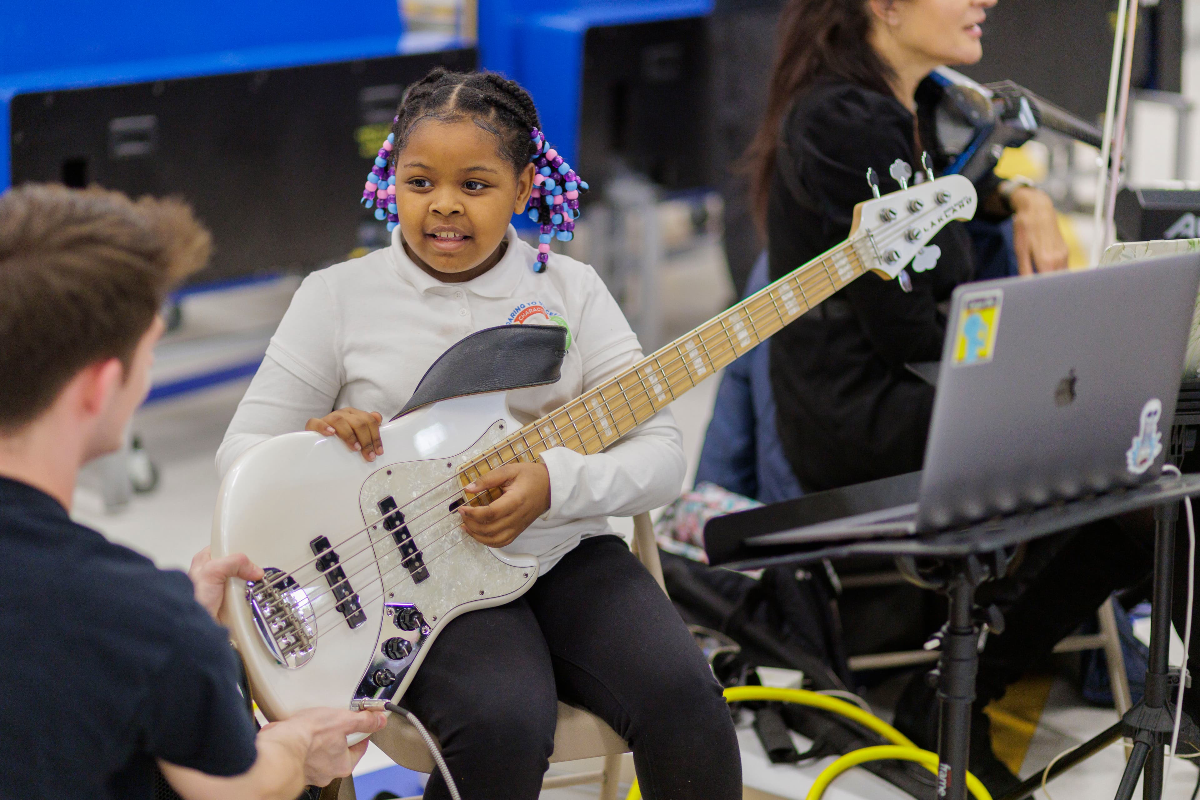 A young student smiles while holding a bass guitar handed to her by one of the performing musicians